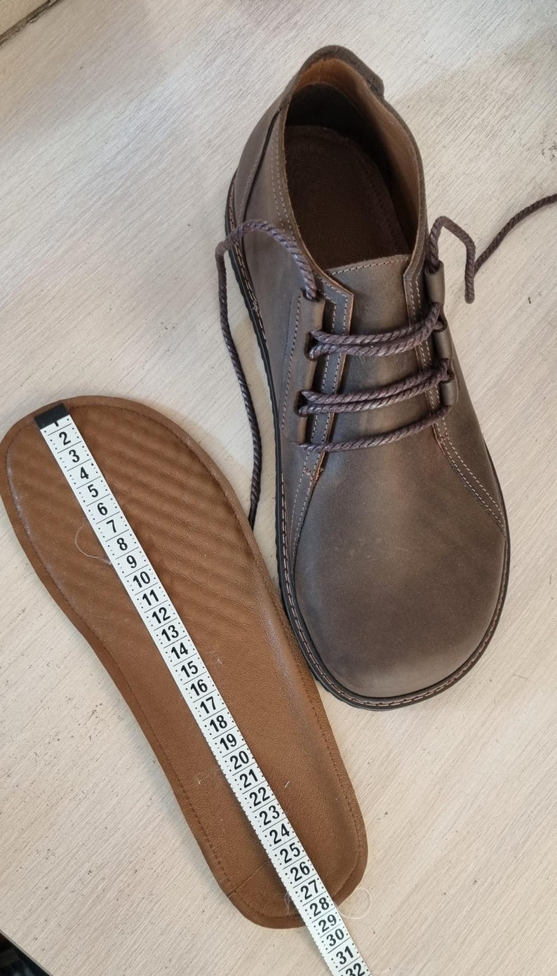 May include: A brown leather shoe with laces and a matching insole. The insole is measured with a ruler, showing a length of approximately 30 cm. The shoe has a rounded toe and a textured appearance.