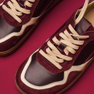 May include: Burgundy and cream-colored sneakers with beige laces. The shoes feature a combination of leather and suede materials. The design includes a cream-colored stripe along the sides and a brown sole. The sneakers are set against a burgundy background.