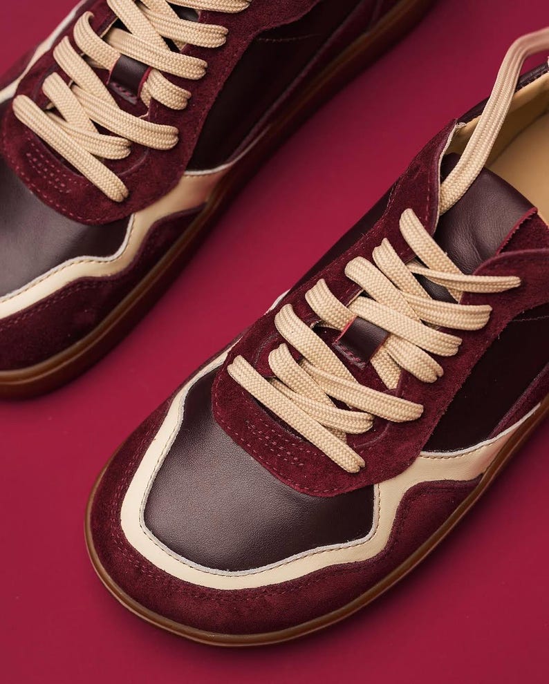 May include: Burgundy and cream-colored sneakers with beige laces. The shoes feature a combination of leather and suede materials. The design includes a cream-colored stripe along the sides and a brown sole. The sneakers are set against a burgundy background.