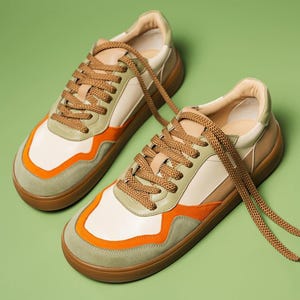 May include: A pair of sneakers with a cream-colored upper, accented with orange and olive green panels. The shoes have brown laces and soles. The sneakers are displayed on a green background.