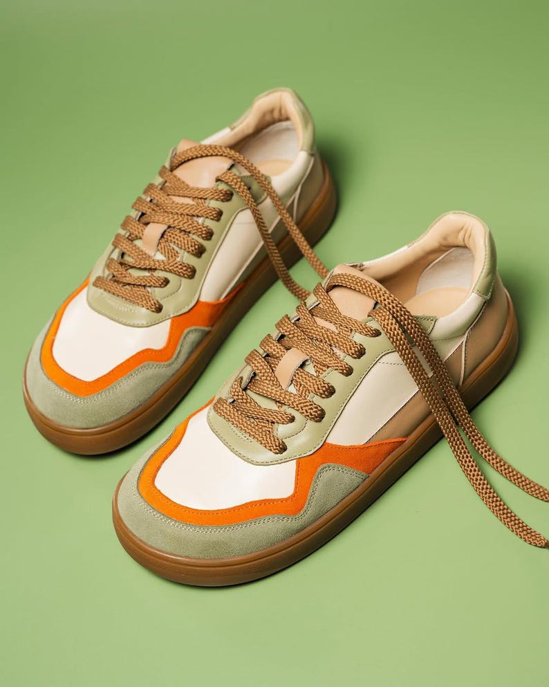 May include: A pair of sneakers with a cream-colored upper, accented with orange and olive green panels. The shoes have brown laces and soles. The sneakers are displayed on a green background.