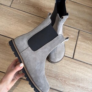 May include: A pair of gray suede Chelsea boots with black elastic side panels and a black lug sole. The boots have a pull tab at the back and a light brown trim around the sole. The boots are displayed on a wooden surface.