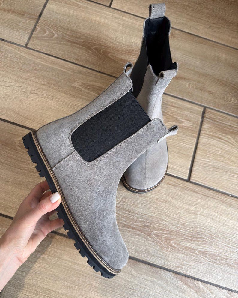 May include: A pair of gray suede Chelsea boots with black elastic side panels and a black lug sole. The boots have a pull tab at the back and a light brown trim around the sole. The boots are displayed on a wooden surface.