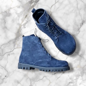 May include: Two blue suede ankle boots with matching laces are displayed on a marble surface. One boot is upright, while the other lies flat, showcasing the lace-up design and textured soles. The boots have a classic, rugged style.