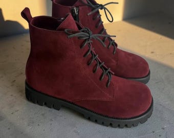 May include: A pair of ankle-high boots in a deep burgundy suede finish. These boots feature black laces, a side zipper closure, a pull tab at the heel, and a thick, black lugged rubber sole with a prominent tread pattern for traction.