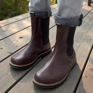 May include: Brown leather Chelsea boots with a rounded toe and a black elastic side panel. The boots have a stitched sole with a light brown trim and are worn with rolled-up gray jeans.