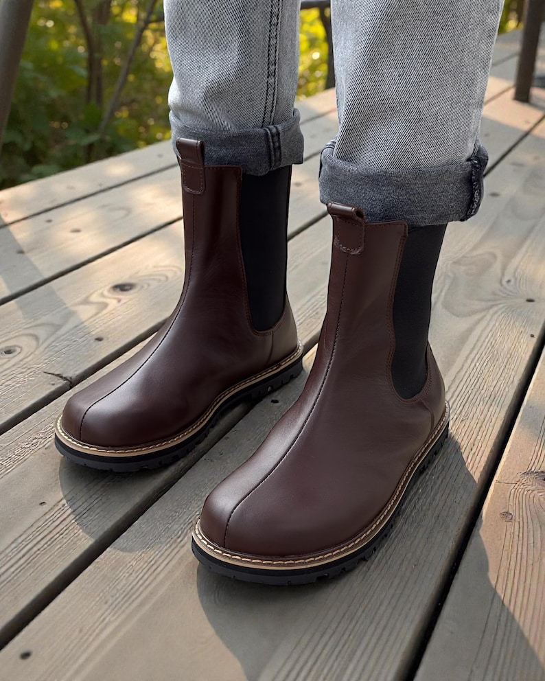 May include: Brown leather Chelsea boots with a rounded toe and a black elastic side panel. The boots have a stitched sole with a light brown trim and are worn with rolled-up gray jeans.