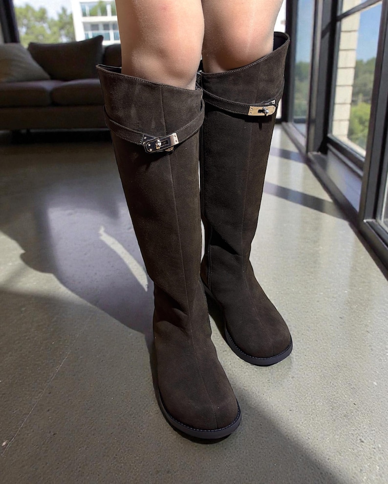 May include: A pair of dark brown suede knee-high boots featuring a rounded toe, a flat sole, and a decorative horizontal strap across the upper shaft secured with a polished silver-tone metal buckle closure on the outer side of each boot.