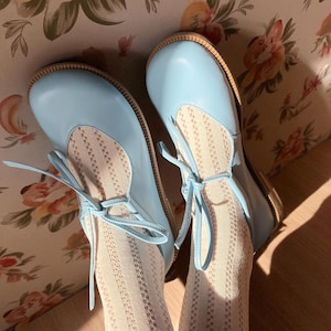 Op de afbeelding: Lichtblauwe Mary Jane-schoenen met strikveters, beige zolen en stoffen inzetstukken met patronen. Gedragen met witte panty's met patronen. De achtergrond is bloemenbehang.