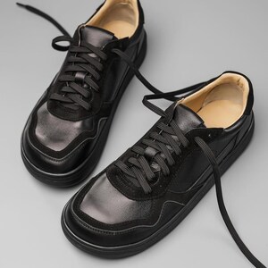 May include: A pair of black leather sneakers with black laces. The shoes have a rounded toe and a thick, black sole. The upper part of the shoe features a combination of smooth and suede-like textures. The interior lining is a light tan color.