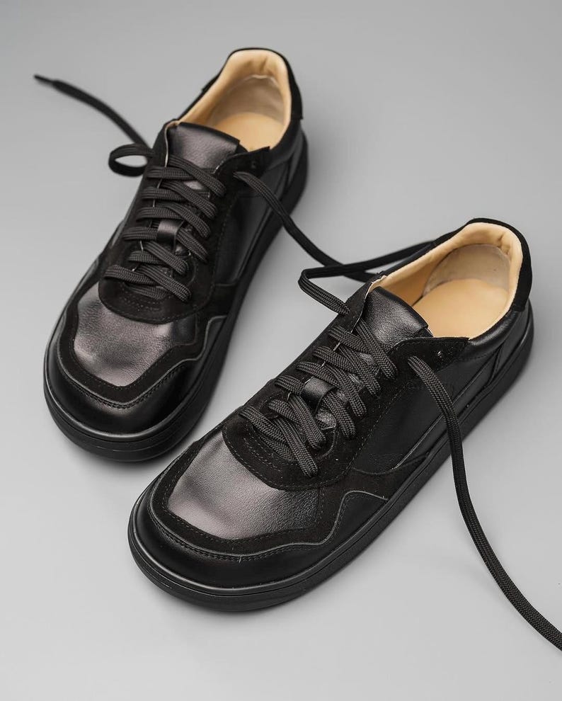 May include: A pair of black leather sneakers with black laces. The shoes have a rounded toe and a thick, black sole. The upper part of the shoe features a combination of smooth and suede-like textures. The interior lining is a light tan color.
