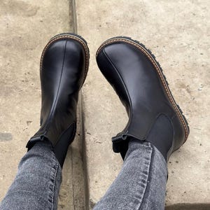 May include: A pair of black leather Chelsea boots with a round toe and a brown stitched sole. The boots have elastic side panels and are worn with gray denim jeans.
