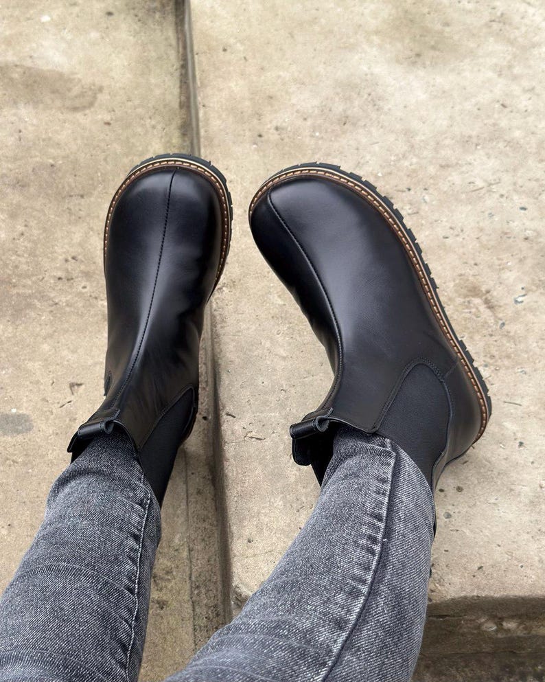 May include: A pair of black leather Chelsea boots with a round toe and a brown stitched sole. The boots have elastic side panels and are worn with gray denim jeans.