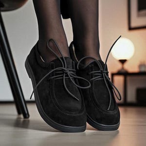 May include: Black suede shoes with lace-up detailing. The shoes have a rounded toe and a flat sole. The image is taken from a low angle, highlighting the shoes. The shoes are a classic design, suitable for various occasions.