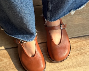 May include: A pair of cognac-colored leather Mary Jane shoes featuring a rounded toe, a deep V-shaped cutout at the vamp, and a thin adjustable strap with a small dark metal buckle across the instep, worn with blue denim jeans.