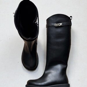 May include: A pair of black knee-high boots featuring a smooth finish, a rounded toe, and a flat sole. Each boot includes a decorative horizontal strap across the upper shaft secured with a silver-toned metal turn-lock clasp and a pull tab at the top.