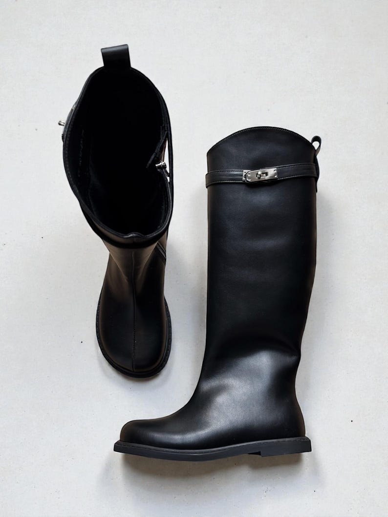May include: A pair of black knee-high boots featuring a smooth finish, a rounded toe, and a flat sole. Each boot includes a decorative horizontal strap across the upper shaft secured with a silver-toned metal turn-lock clasp and a pull tab at the top.