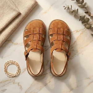 May include: A pair of brown suede sandals with a caged design. The sandals have a buckle closure and a cream-colored insole. The shoes are displayed on a marble surface, with a folded beige fabric and a beaded bracelet nearby.