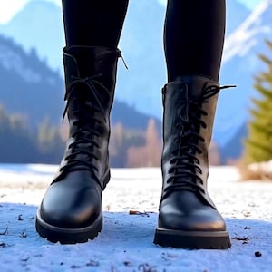 May include: A pair of black leather combat boots with laces and a lug sole. The boots are worn with black leggings, and the background features a snowy landscape with mountains and trees.