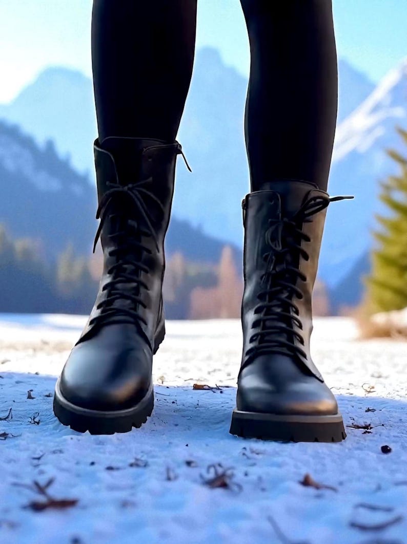 May include: A pair of black leather combat boots with laces and a lug sole. The boots are worn with black leggings, and the background features a snowy landscape with mountains and trees.