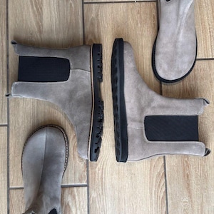 May include: A collection of gray suede Chelsea boots with black elastic side panels and black soles. The boots are arranged on a light brown wooden floor, showcasing their design and construction. The boots are a neutral color, suitable for various outfits.