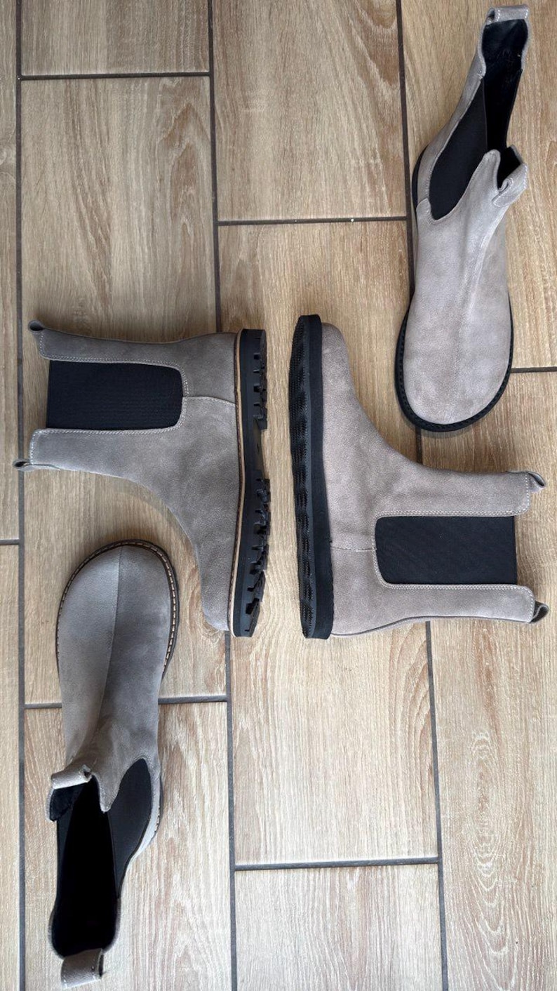 May include: A collection of gray suede Chelsea boots with black elastic side panels and black soles. The boots are arranged on a light brown wooden floor, showcasing their design and construction. The boots are a neutral color, suitable for various outfits.