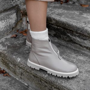 May include: Gray leather boots with a white sole and a front zipper. The boots have a round toe and are worn with white socks. The boots are on a stone surface.