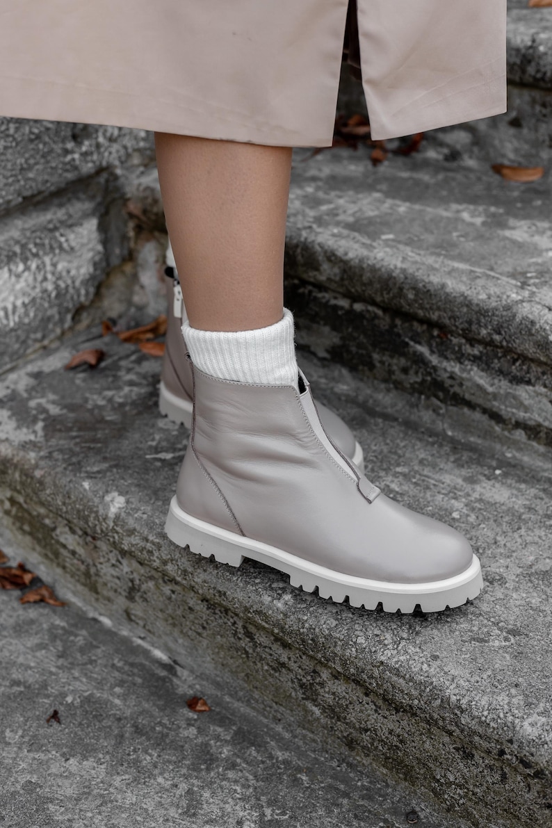 May include: Gray leather boots with a white sole and a front zipper. The boots have a round toe and are worn with white socks. The boots are on a stone surface.