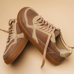 May include: A pair of brown and tan sneakers with matching laces. The shoes have a low-profile design with a rounded toe and a rubber sole. The sneakers are made of leather and suede materials.