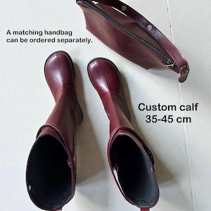 May include: A pair of dark red boots with a rounded toe and a matching zippered handbag. The boots feature a strap detail and a custom calf measurement of 35-45 cm. The text states: A matching handbag can be ordered separately. Custom calf 35-45 cm.