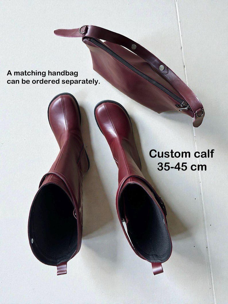 May include: A pair of dark red boots with a rounded toe and a matching zippered handbag. The boots feature a strap detail and a custom calf measurement of 35-45 cm. The text states: A matching handbag can be ordered separately. Custom calf 35-45 cm.