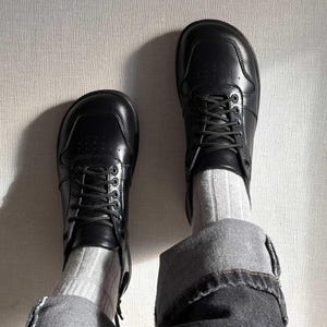 May include: A pair of black leather sneakers with laces, worn with gray socks and denim jeans. The sneakers have a classic design with a rounded toe and a low profile. The jeans are cuffed at the ankles.