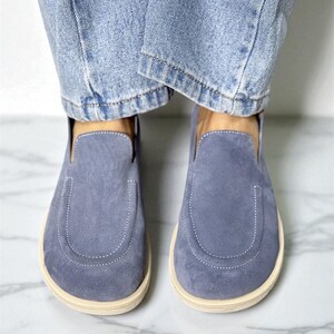 Op de afbeelding: Een paar blauwe su&egrave;de instappers met een afgeronde neus en een cr&egrave;mekleurige zool. De schoenen worden gedragen met lichtblauwe spijkerbroeken. De schoenen hebben een gestikt detail rond de teen.