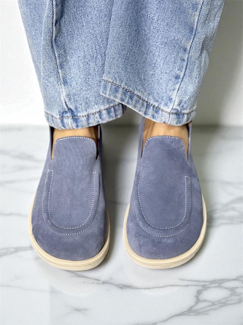 Op de afbeelding: Een paar blauwe su&egrave;de instappers met een afgeronde neus en een cr&egrave;mekleurige zool. De schoenen worden gedragen met lichtblauwe spijkerbroeken. De schoenen hebben een gestikt detail rond de teen.
