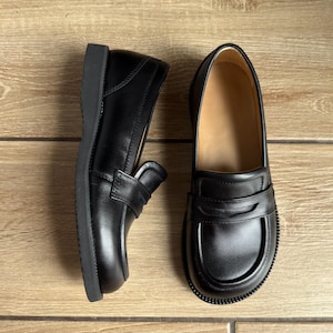 Puede incluir: Un par de mocasines de cuero negro con una cl&aacute;sica tira con ranura sobre el empeine, punta redondeada y una suela de goma negra gruesa y duradera. El interior de los zapatos est&aacute; forrado con un material de color tostado claro que contrasta.