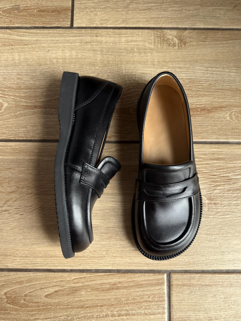 Puede incluir: Un par de mocasines de cuero negro con una cl&aacute;sica tira con ranura sobre el empeine, punta redondeada y una suela de goma negra gruesa y duradera. El interior de los zapatos est&aacute; forrado con un material de color tostado claro que contrasta.