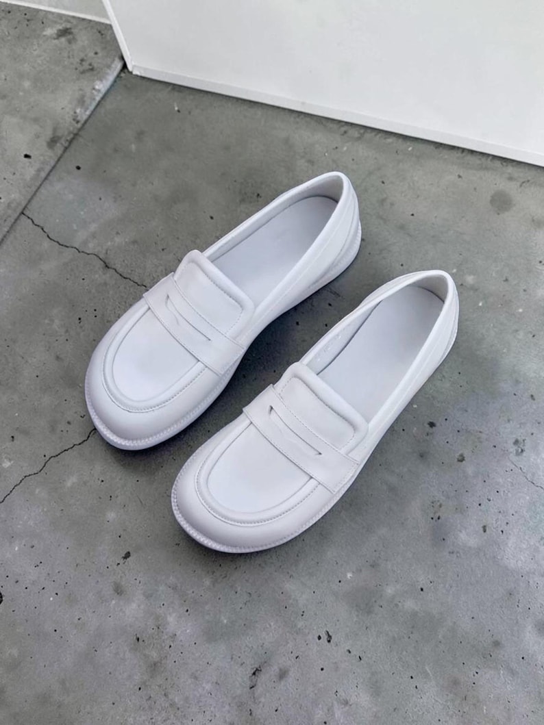 Puede incluir: Un par de mocasines blancos lisos sin cordones, con punta redondeada, una tira cl&aacute;sica con ranura sobre el empeine y un acabado liso y limpio. Estos zapatos tienen un dise&ntilde;o de perfil bajo con forro interior blanco y una suela resistente.