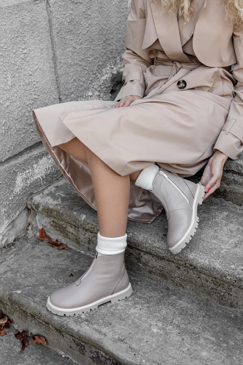 May include: Light gray leather boots with white soles and a zipper closure. The boots have a chunky, lugged sole and are paired with white knit socks. The boots are part of a fall fashion ensemble.