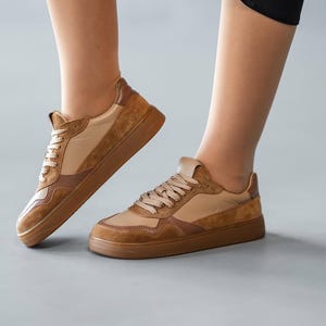 May include: Tan and brown sneakers with a low profile. The shoes feature a combination of smooth and suede-like materials, with a light tan base and darker brown accents. The laces are a matching tan color.