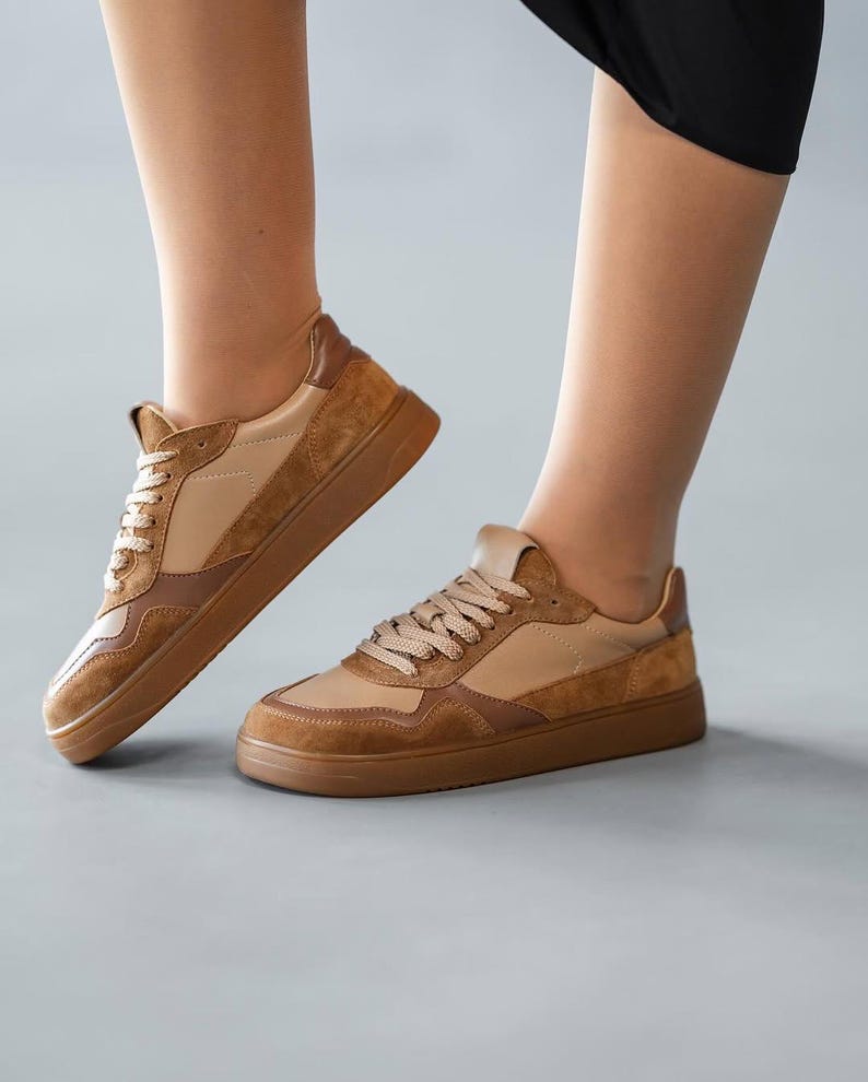 May include: Tan and brown sneakers with a low profile. The shoes feature a combination of smooth and suede-like materials, with a light tan base and darker brown accents. The laces are a matching tan color.