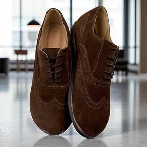 Könnte beinhalten: Ein Paar dunkelbraune Wildleder-Oxford-Schuhe mit klassischem Flügelkappen-Design, dekorativen Brogue-Perforationen entlang der Nähte, passenden braunen Schnürsenkeln und einem hellen Innenfutter, die in aufrechter Position präsentiert werden.