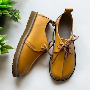 May include: Mustard-yellow leather shoes with brown laces. The shoes have a rounded toe and a thick, brown sole. The laces are tied in a bow. The shoes are set against a white background. The shoes are approximately 25 cm long.