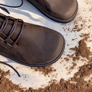 May include: Close-up of two brown leather boots with dark brown laces. The boots have a rounded toe and a stitched sole. The background is a light surface with scattered brown soil.