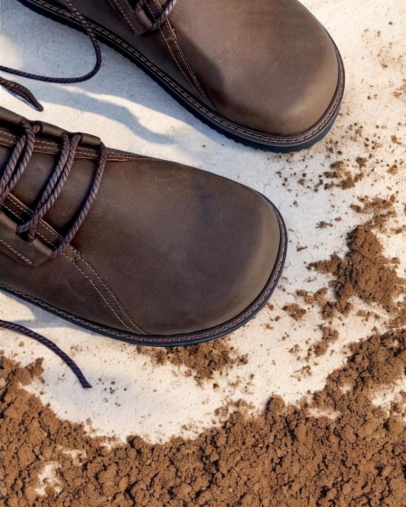 May include: Close-up of two brown leather boots with dark brown laces. The boots have a rounded toe and a stitched sole. The background is a light surface with scattered brown soil.