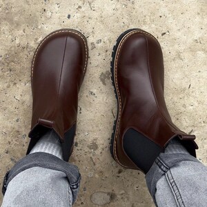 May include: A pair of brown leather Chelsea boots with a rounded toe and a black elastic side panel. The boots have a thick, textured sole and a stitched welt. The boots are worn with gray socks and denim jeans.