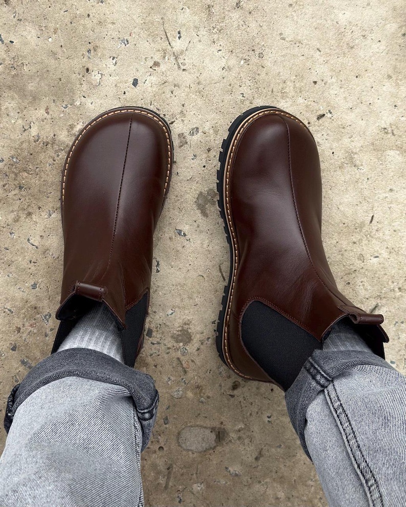 May include: A pair of brown leather Chelsea boots with a rounded toe and a black elastic side panel. The boots have a thick, textured sole and a stitched welt. The boots are worn with gray socks and denim jeans.