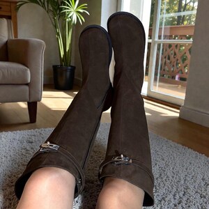 May include: A pair of dark brown suede knee-high boots featuring a rounded toe, a flat sole, and a decorative strap across the ankle secured with a silver-toned metal buckle hardware detail. The boots have a side zipper closure and a smooth finish.