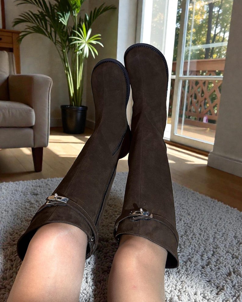 May include: A pair of dark brown suede knee-high boots featuring a rounded toe, a flat sole, and a decorative strap across the ankle secured with a silver-toned metal buckle hardware detail. The boots have a side zipper closure and a smooth finish.