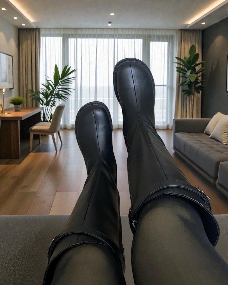 May include: A pair of black knee-high boots featuring a smooth leather-like finish, a rounded toe box with a central vertical seam, and a decorative strap detail encircling the upper ankle area with a metallic buckle closure on the outer side.
