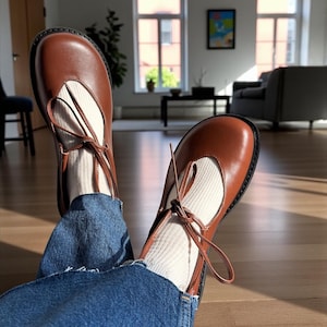 Op de afbeelding: Bruine leren Mary Jane-schoenen met een ronde neus en enkelbandjes. De schoenen worden gedragen met witte sokken en een blauwe spijkerbroek. De schoenen zijn ongeveer 23 cm lang.